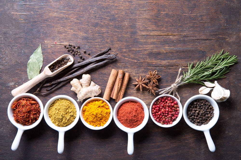 Bunte Gewürze und Kräuter in weißen Schalen auf einem Holztisch, darunter Safran, Kurkuma, Paprika, Pfeffer und Zimtstangen.