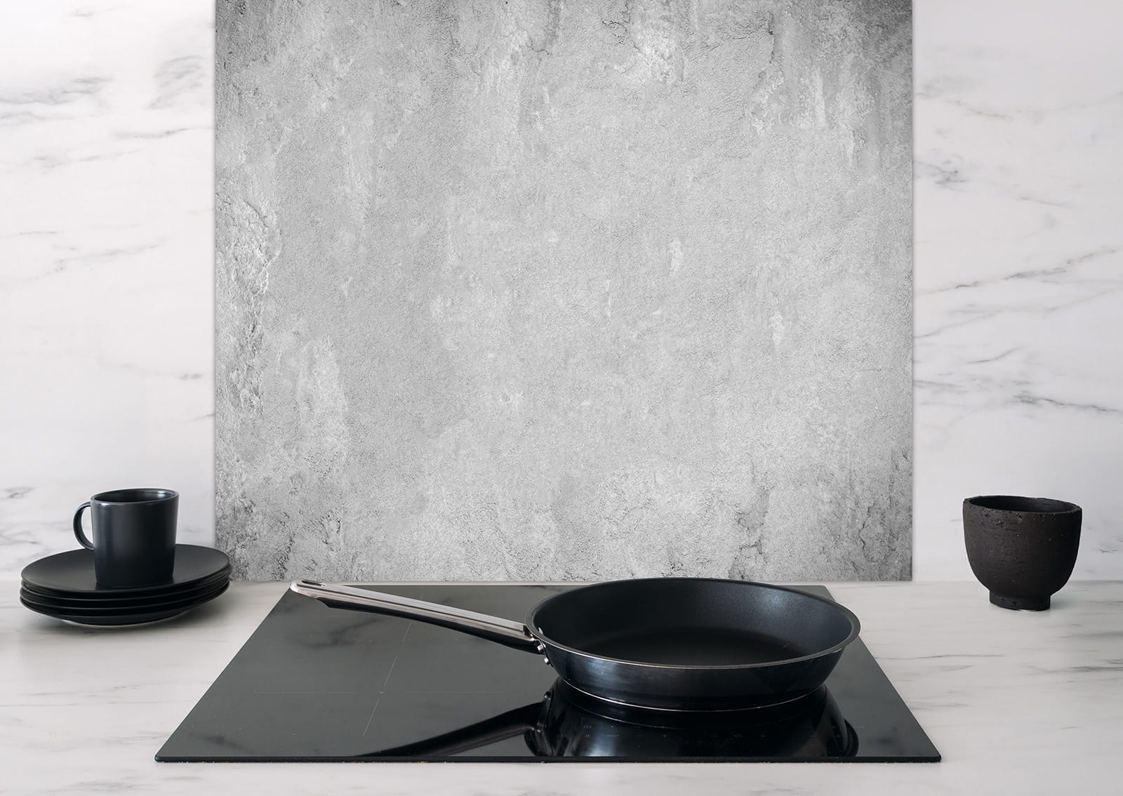 Spritzschutz Heller Beton von ENTDECKER, graue Betontextur im Hintergrund, schwarze Pfanne auf Kochfeld, Geschirr in Schwarz.