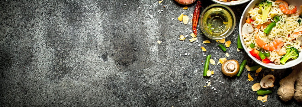 Schüssel mit Nudeln, Gemüse und Garnelen auf einem dunklen Tisch, umgeben von Gewürzen und Zutaten.