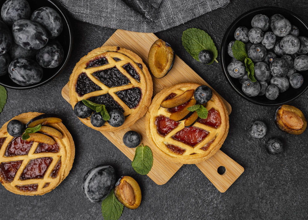 Drei kleine Kuchen mit Marmelade und frischen Beeren auf einem Holzbrett, umgeben von Blaubeeren und Pflaumenhälften.